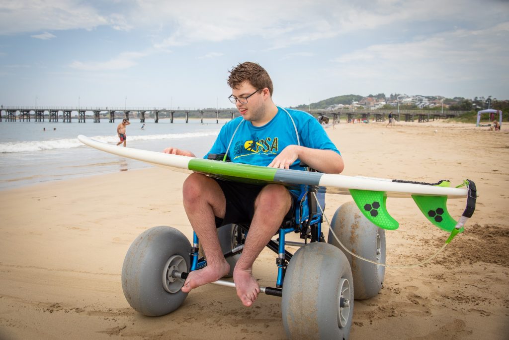 Mid shot of Brendan Harvey using a beach wheelchair and holding a surfboard at Jetty Beach