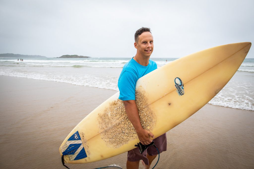 Mid shot of Jordy Davis holding a surfboard with the ocean behind him