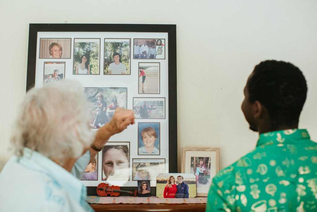 A young man and an elderly woman look at framed family photos on a wall.