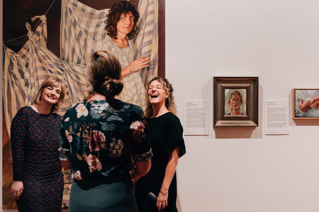 Two women smile in front of a large portrait at Coffs Harbour Regional Gallery