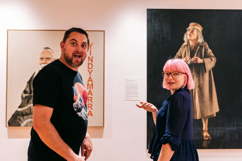 A man and a woman with pink hair with large portraits in the background