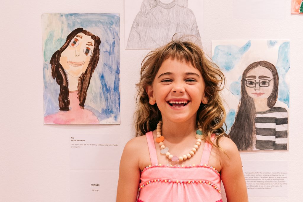 A little girl smiling with a drawing she has done in the background.
