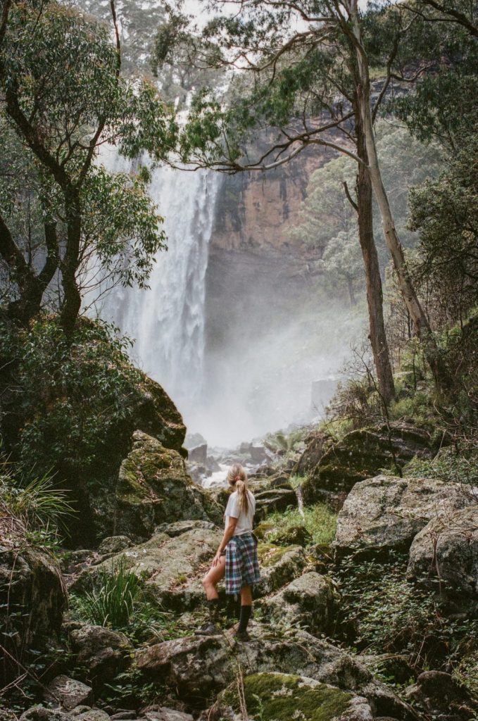 A woman stands amongst rocks facing a waterfall 