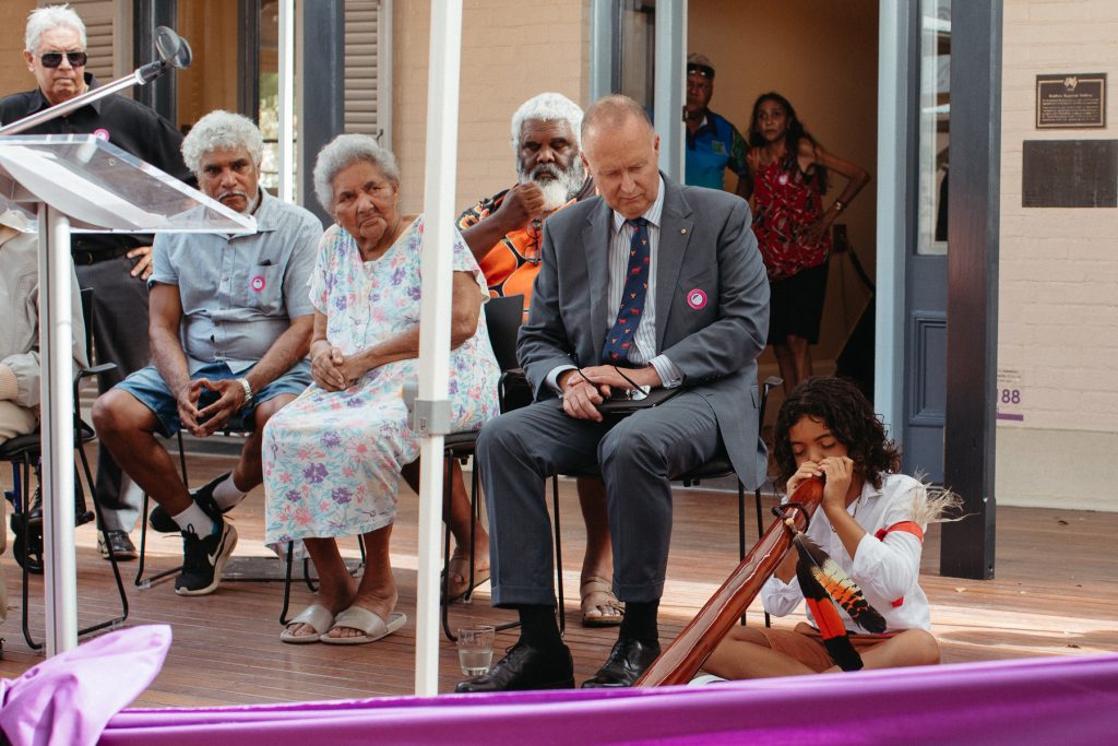 A young Aboriginal boy plays the didgeridoo in front of a seated audience