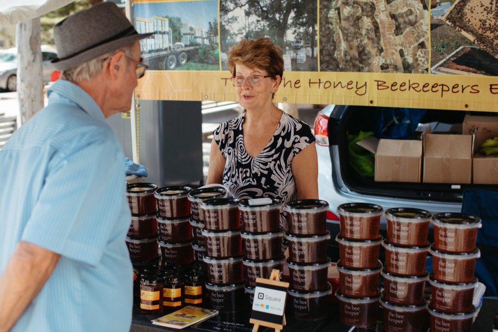 A woman speaks to a man next to stacks of honey in plastic tubs.