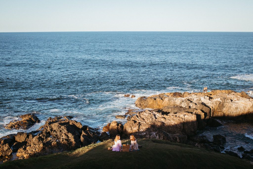 Wide landscape shot of two young women having a picnic on a headland with the ocean in the background