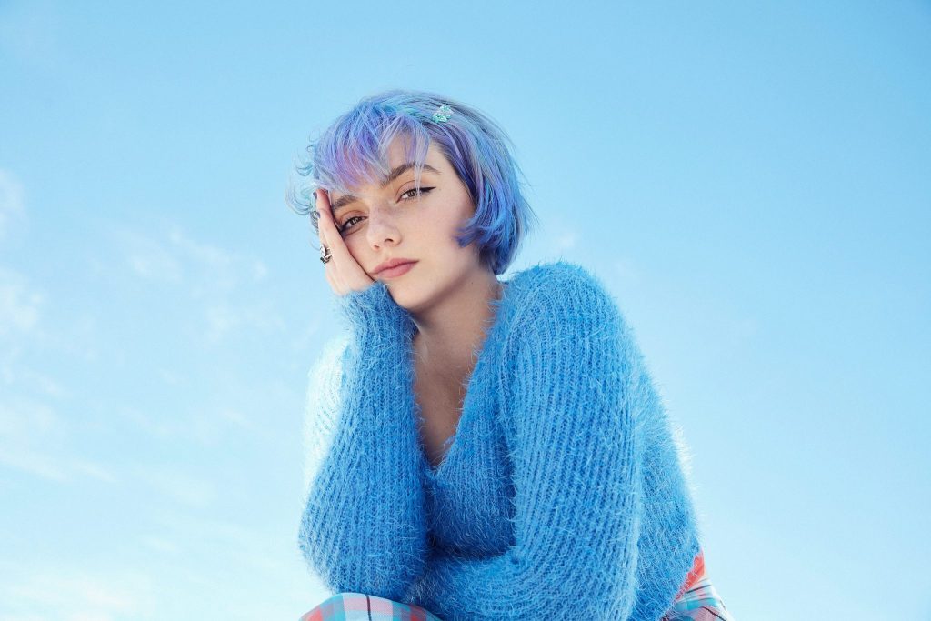 A young woman with blue hair and a blue knit jumper looking at the camera