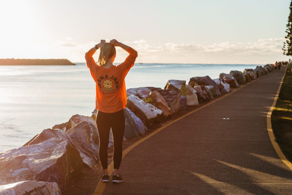 A woman wearing an orange long sleeve shirt and holding yoga mat walks along a boardwalk at sunrise