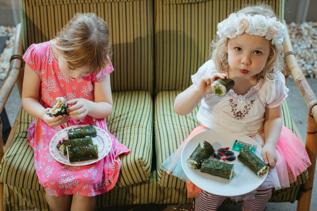 Two little girls eat sushi on a green couch 